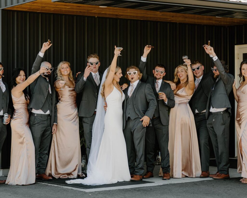 Bridal party toasts to being newlyweds with heart-shaped sunglasses, champagne coups raised in the air, and cigars in their mouths