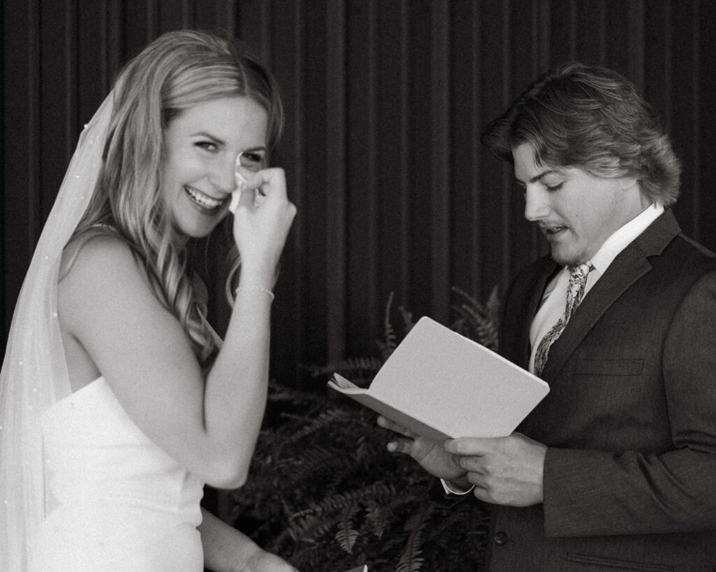 Bride smiles at camera as she cries and groom reads her his vows