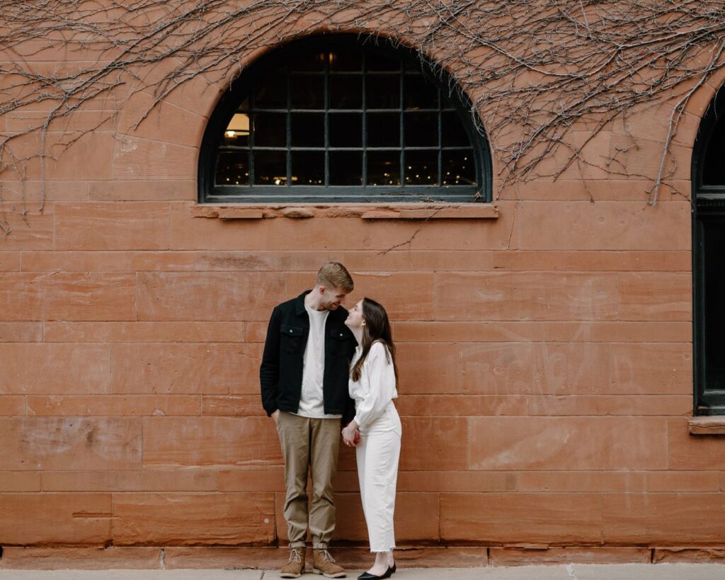 Engaged couple grins at each other in front of brownstone architecture and teal windows. 