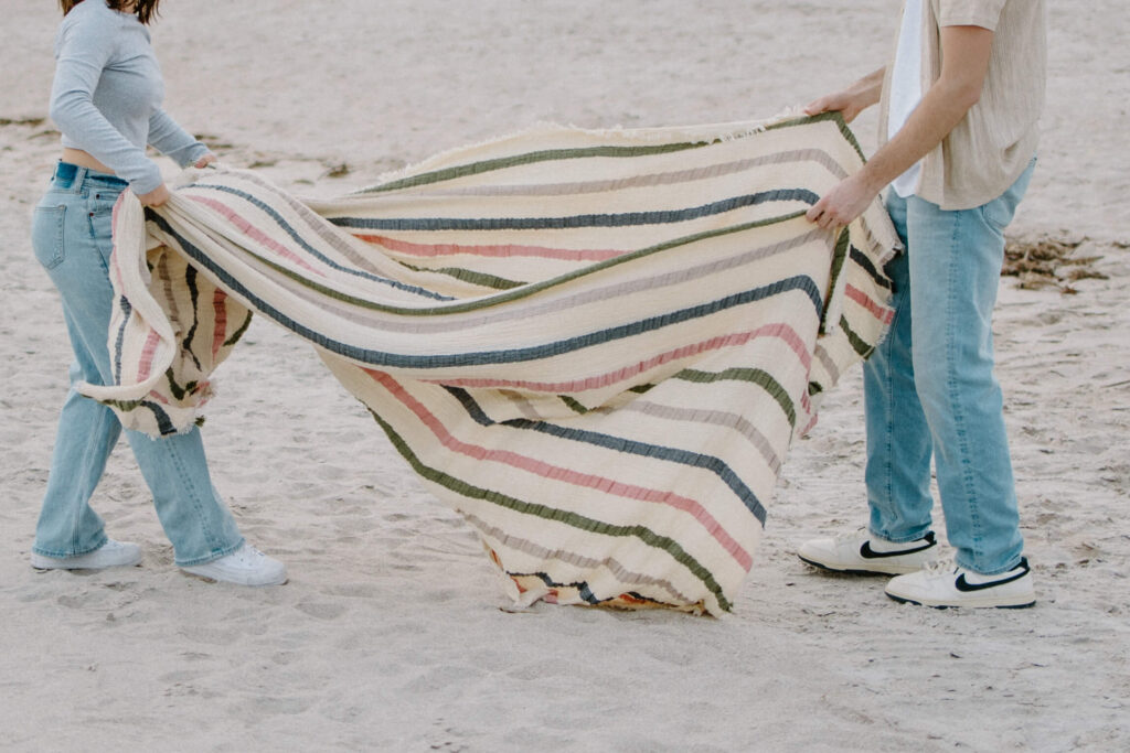 Couple spreads striped blanket on the sandy beach in Sanibel Island adventure engagement photoshoot. 