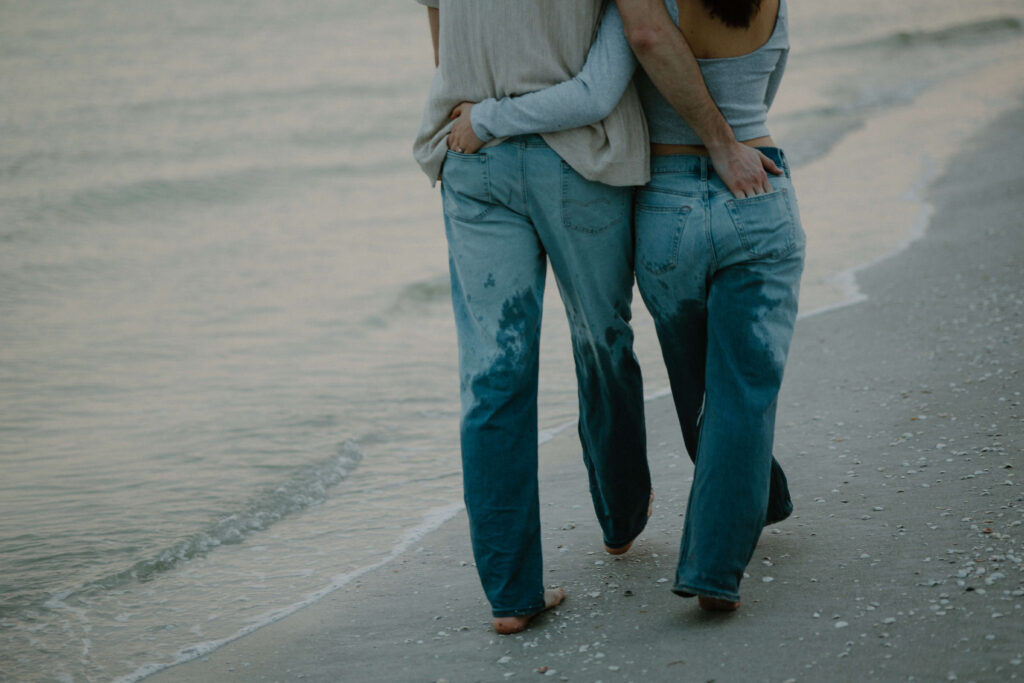 Engaged couple walks up and down the beach with soaked jeans and their hands in each other's back pockets.