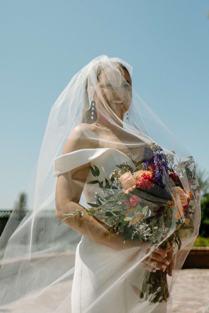 Bride smiles with veil over her face in direct sunlight