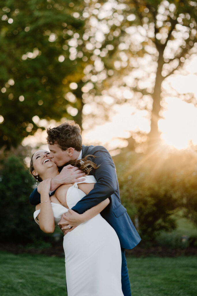 Groom hugs and kisses bride as golden hour sunlight cascades behind them