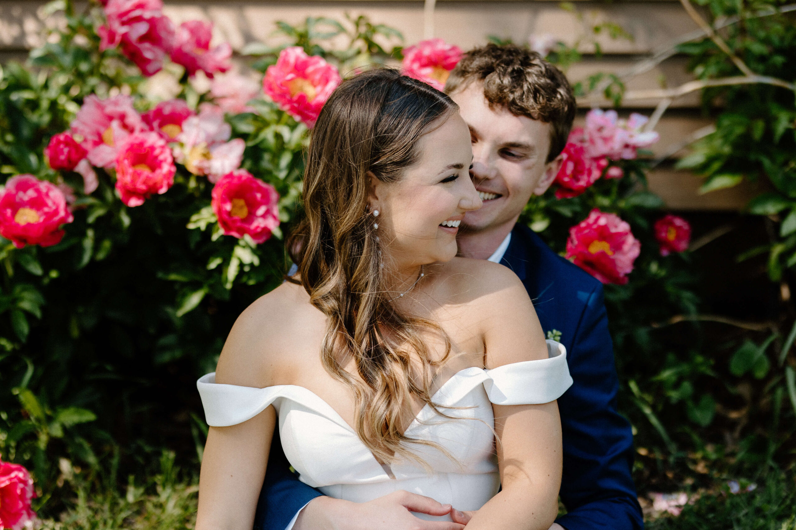 Documentary-style photographer captures couple sitting in front of bright pink peonies on their wedding day