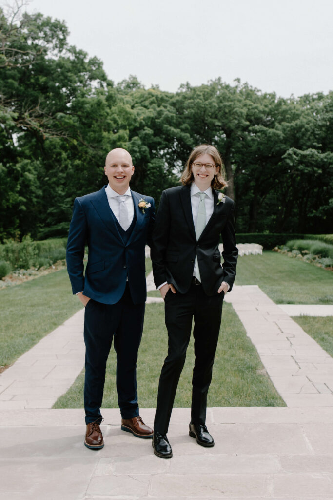 Groom and best man smile with hands in their pockets in garden wedding