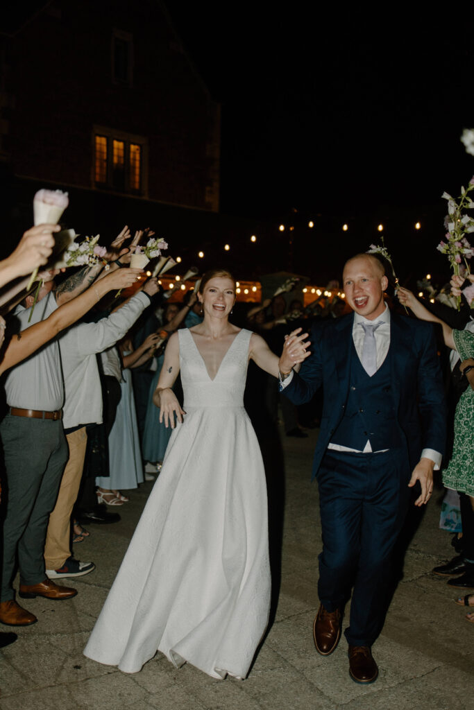 Bride and groom make their grand exit as husband and wife and garden wedding guests hold flowers up in the air