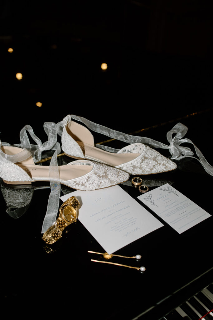 Lace bridal shoes, watch, pearl hair clips, and wedding invitation suite sit on Yamaha piano bench