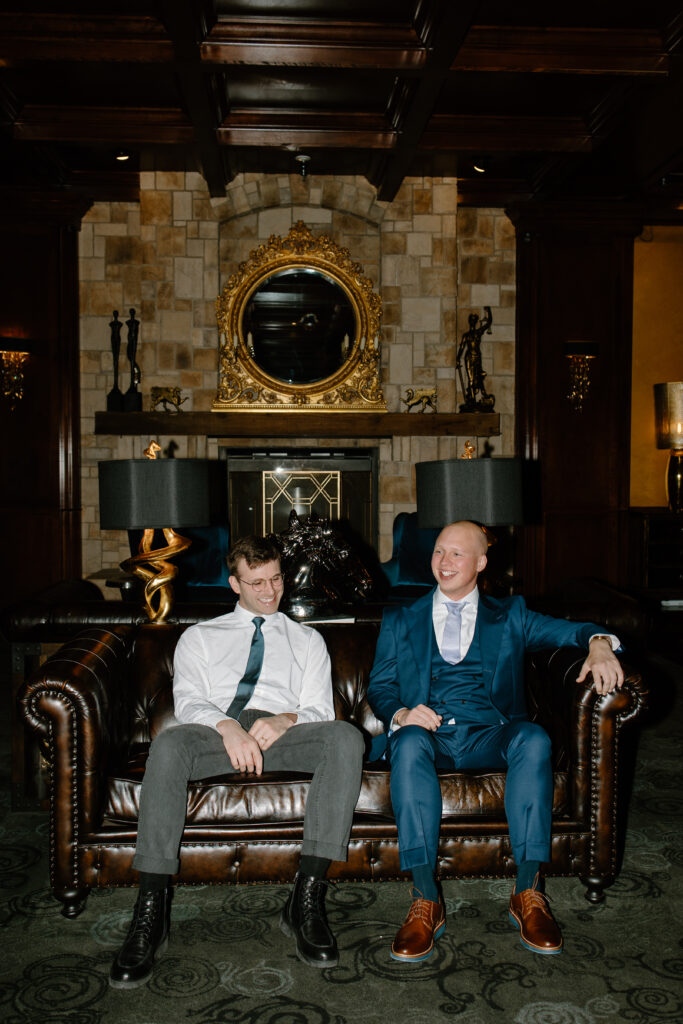 Groom and his best friend laugh while they sit on a luxury leather couch