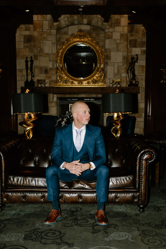 Groom sits on leather couch in navy Bespoke Custom Clothing suit