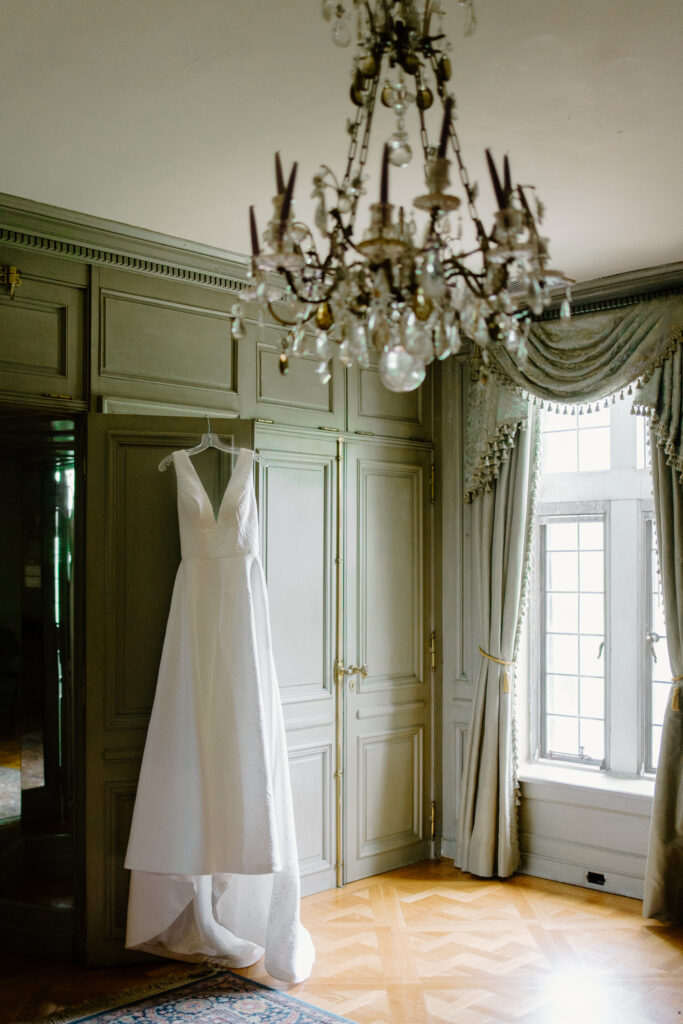 Wedding gown hangs in the closet of an elegant French-style room with sage green walls and a chandelier