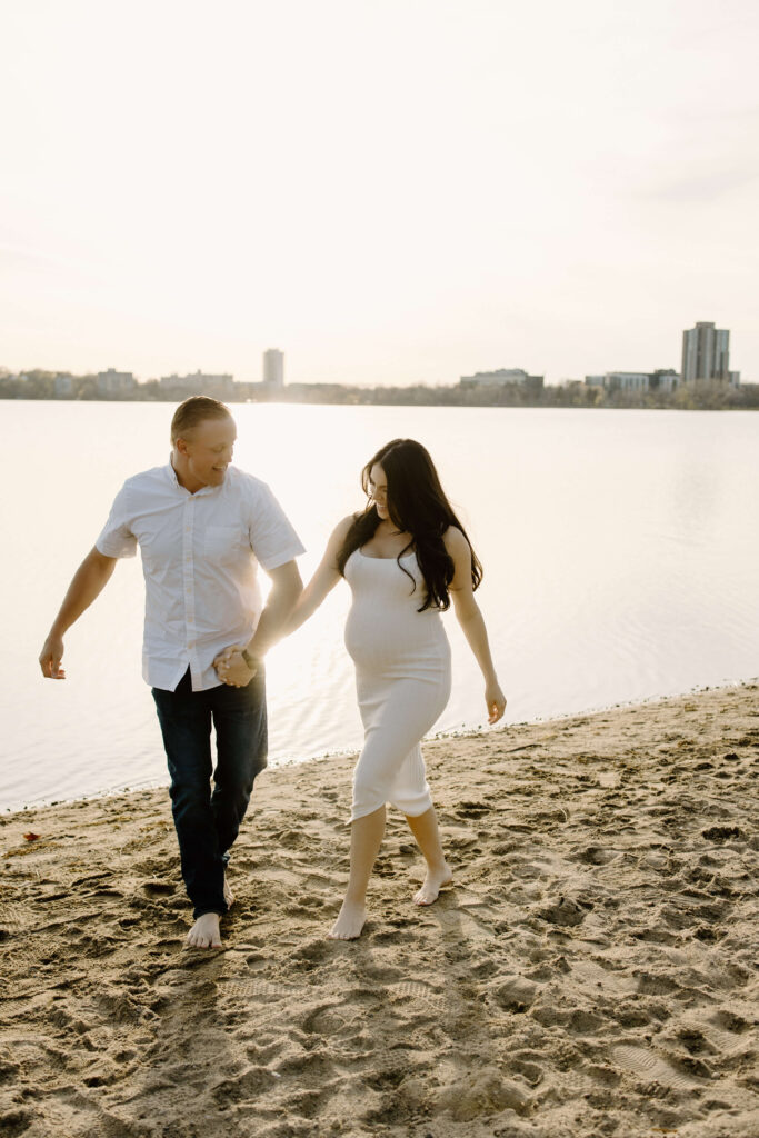 Man holds pregnant woman's hand as she walks in the sand along Bde Mka Ska.